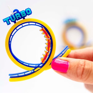 Pu&ograve; includere: Un paio di orecchini a forma di montagne russe blu e gialle con la scritta "TURBO" in lettere blu. Le montagne russe sono in plastica e hanno vagoni rossi e gialli.