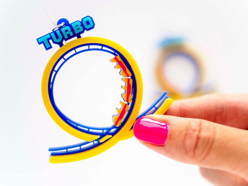 Pu&ograve; includere: Un paio di orecchini a forma di montagne russe blu e gialle con la scritta "TURBO" in lettere blu. Le montagne russe sono in plastica e hanno vagoni rossi e gialli.