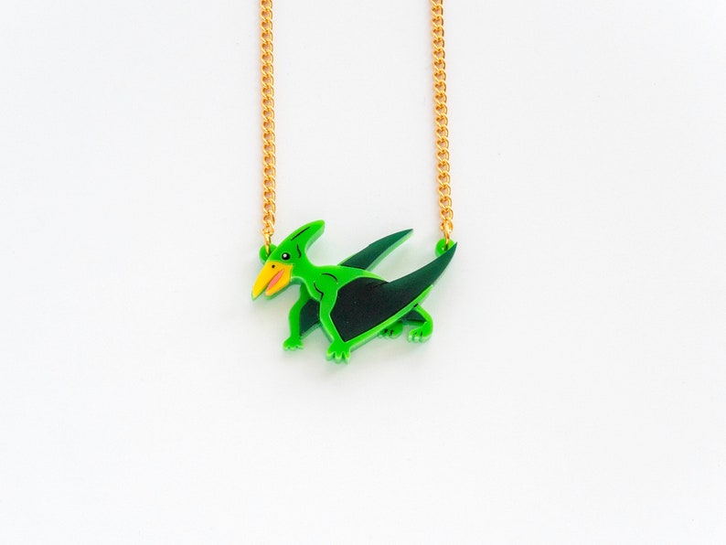 May include: A green and black pterosaur pendant necklace with a gold chain. The pterosaur is depicted with a yellow beak and black eyes.