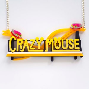 May include: A yellow and black necklace with a silver chain. The necklace features a sign that says "CRAZY MOUSE" with a yellow roller coaster track and two pink and white mouse cars.