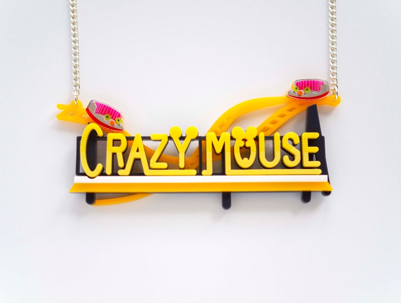 May include: A yellow and black necklace with a silver chain. The necklace features a sign that says "CRAZY MOUSE" with a yellow roller coaster track and two pink and white mouse cars.