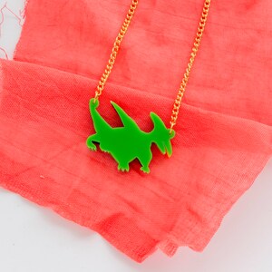 May include: A green dinosaur pendant necklace on a gold chain. The necklace is lying on a pink fabric background.