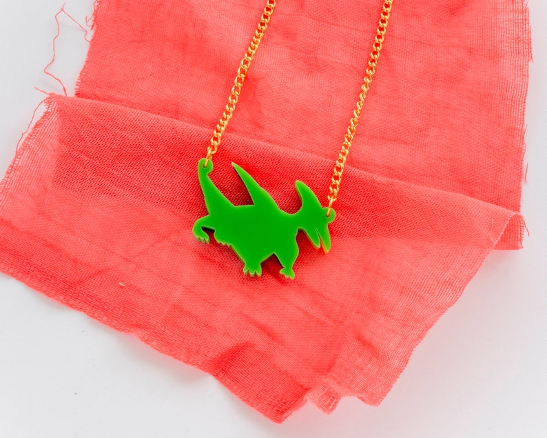 May include: A green dinosaur pendant necklace on a gold chain. The necklace is lying on a pink fabric background.