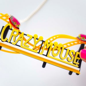May include: A yellow and black necklace with a roller coaster design and the words "Crazy Mouse". The roller coaster has two small pink and yellow cars.