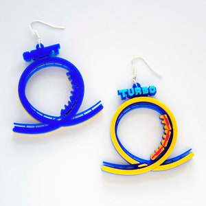 Pu&ograve; includere: Un paio di orecchini a forma di montagne russe blu e gialli. Un orecchino ha la parola "TURBO" in lettere blu.