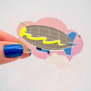 May include: A quirky brooch depicting a grey airship with blue fins and a yellow detail, set against a pink cloud-shaped backdrop. The airship design has silver lines. A small, iridescent cloud is also present.