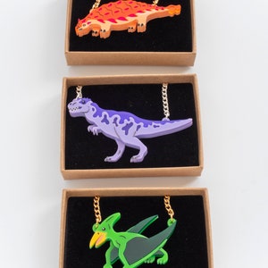 May include: Three necklaces featuring dinosaur and pterosaur pendants. The first necklace has an orange and red ankylosaur pendant. The second necklace has a purple Tyrannosaurus Rex pendant. The third necklace has a green pterodactyl pendant.