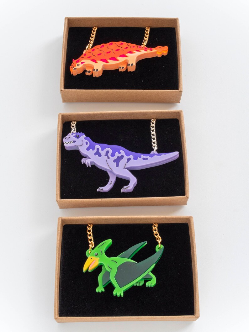 May include: Three necklaces featuring dinosaur and pterosaur pendants. The first necklace has an orange and red ankylosaur pendant. The second necklace has a purple Tyrannosaurus Rex pendant. The third necklace has a green pterodactyl pendant.
