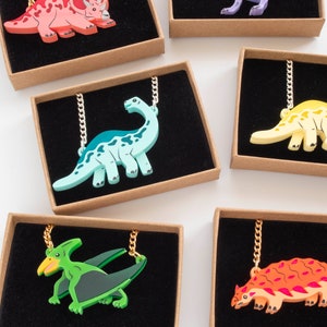 May include: Six different dinosaur necklaces in individual boxes. The necklaces feature a variety of dinosaurs, including a triceratops, a stegosaurus, a pterodactyl, and a T-Rex. The dinosaurs are made of colorful plastic and are attached to gold chains.