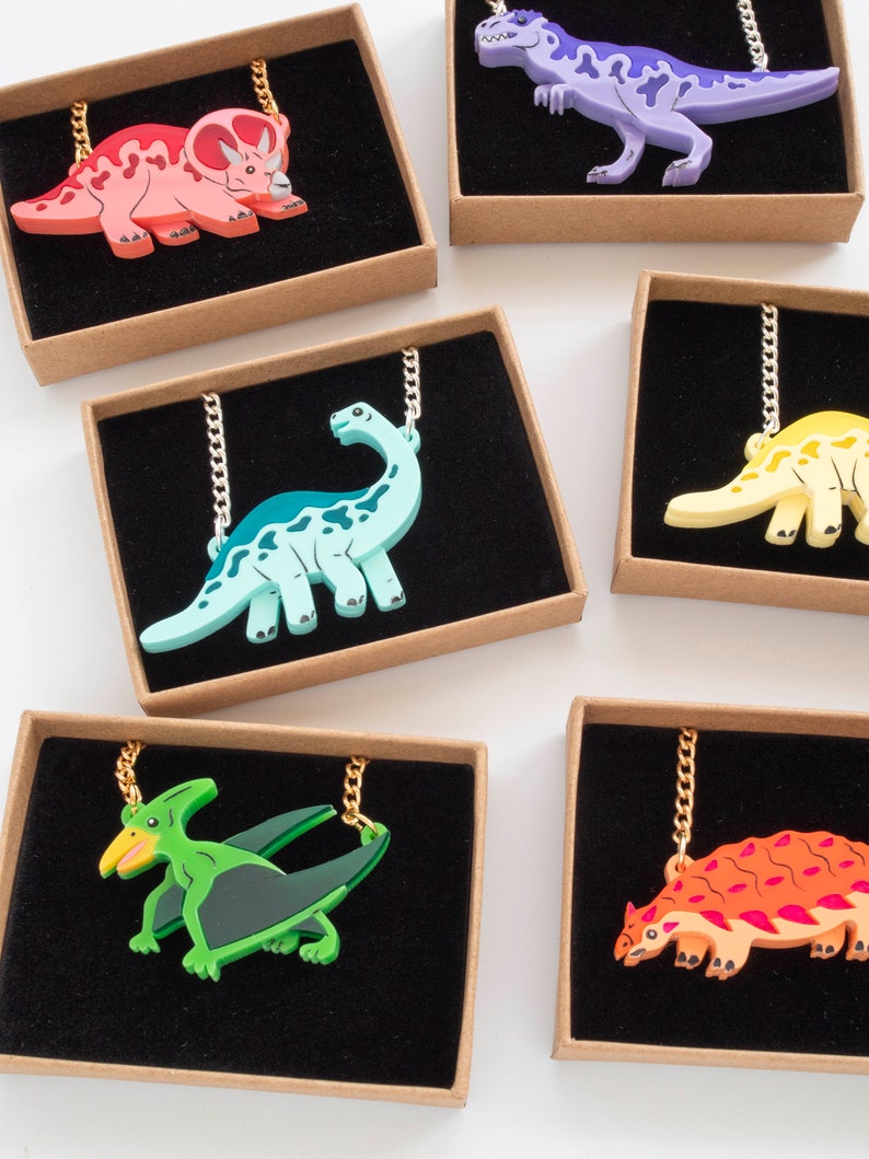 May include: Six different dinosaur necklaces in individual boxes. The necklaces feature a variety of dinosaurs, including a triceratops, a stegosaurus, a pterodactyl, and a T-Rex. The dinosaurs are made of colorful plastic and are attached to gold chains.