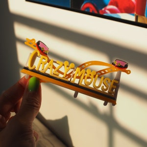 May include: A yellow and black plastic model of a roller coaster with the words "Crazy Mouse" printed on it. The model is held in a hand.