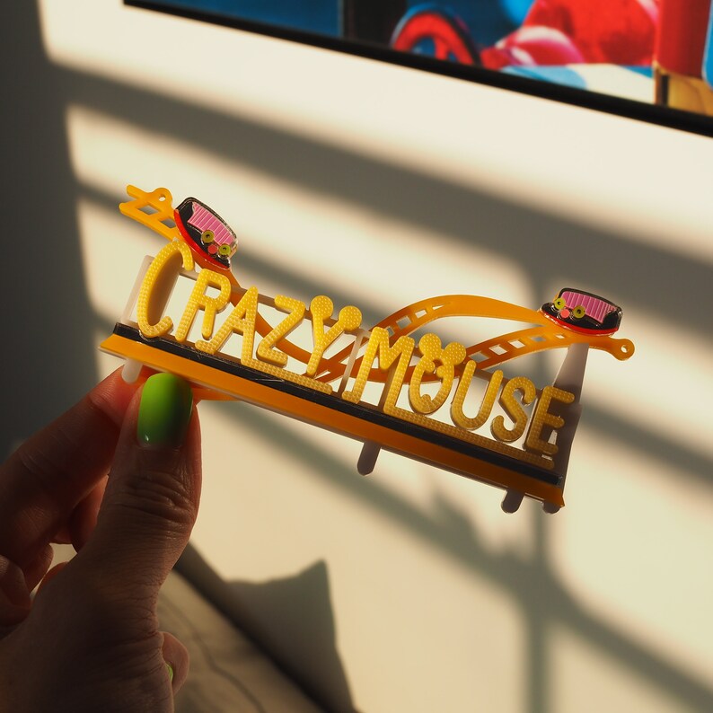 May include: A yellow and black plastic model of a roller coaster with the words "Crazy Mouse" printed on it. The model is held in a hand.