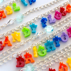 Puede incluir: Un brazalete de cadena plateada con abalorios de letras de colores que deletrean las palabras "APPLE", "CANDY", "BRIGHT", "FLOW" y "LOVE".