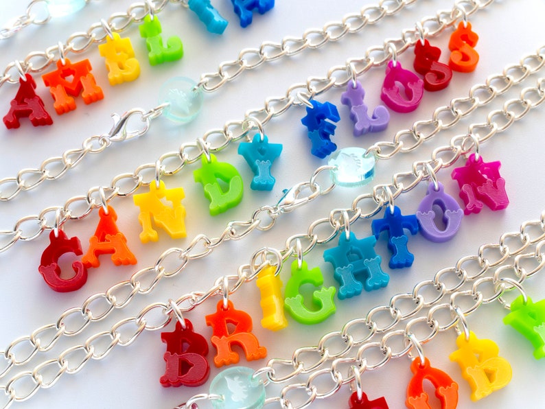 Puede incluir: Un brazalete de cadena plateada con abalorios de letras de colores que deletrean las palabras "APPLE", "CANDY", "BRIGHT", "FLOW" y "LOVE".