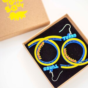 Pu&ograve; includere: Un paio di orecchini a forma di montagne russe blu e gialli con la scritta "TURBO" su ogni orecchino. Gli orecchini sono in una piccola scatola marrone con interno nero.