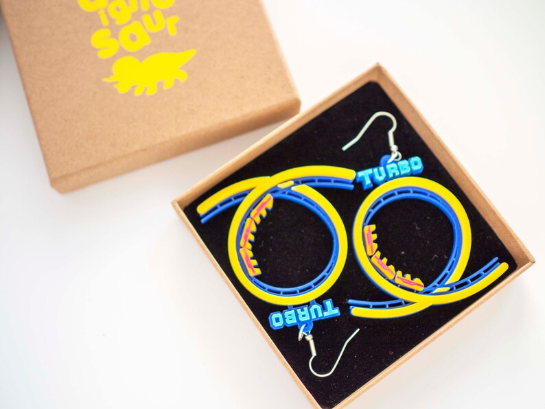 Pu&ograve; includere: Un paio di orecchini a forma di montagne russe blu e gialli con la scritta "TURBO" su ogni orecchino. Gli orecchini sono in una piccola scatola marrone con interno nero.