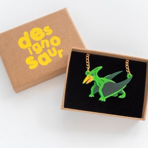 May include: A green and black pterodactyl pendant necklace with a gold chain in a brown box with the text "designosaur" printed in yellow.
