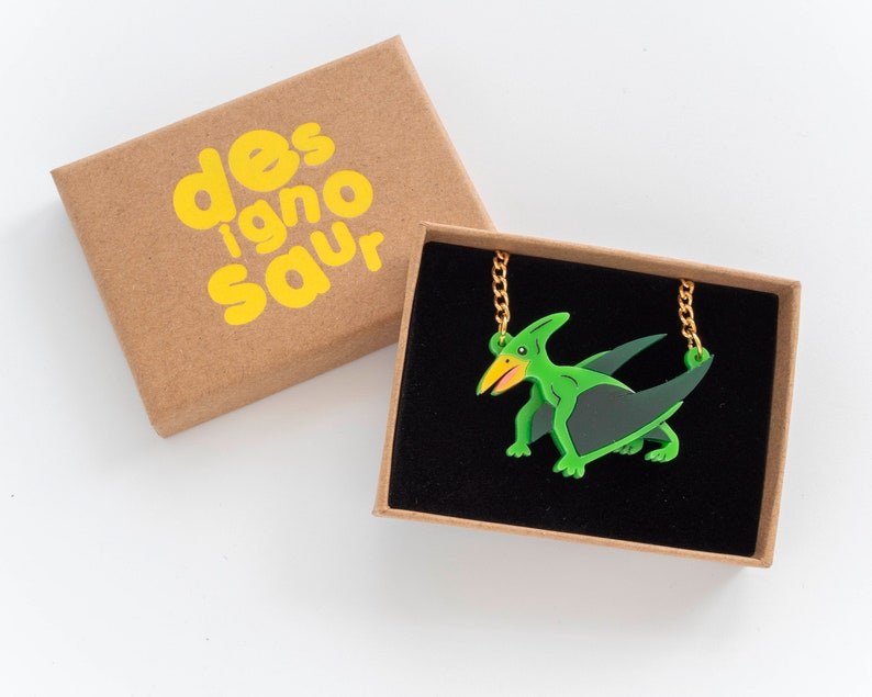 May include: A green and black pterodactyl pendant necklace with a gold chain in a brown box with the text "designosaur" printed in yellow.