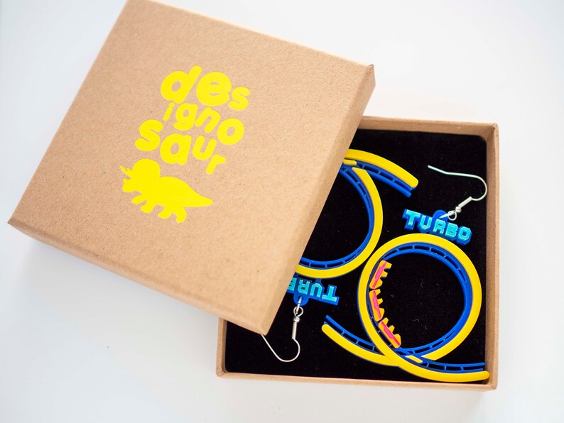 Pu&ograve; includere: Un paio di orecchini a forma di montagne russe gialli e blu con la scritta "TURBO" su di essi. Gli orecchini sono in una piccola scatola marrone con un logo di dinosauro giallo che dice "des'igno'saur".
