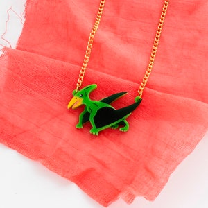 May include: A gold chain necklace with a green and black pterodactyl pendant. The pendant is made of wood and has a detailed design.