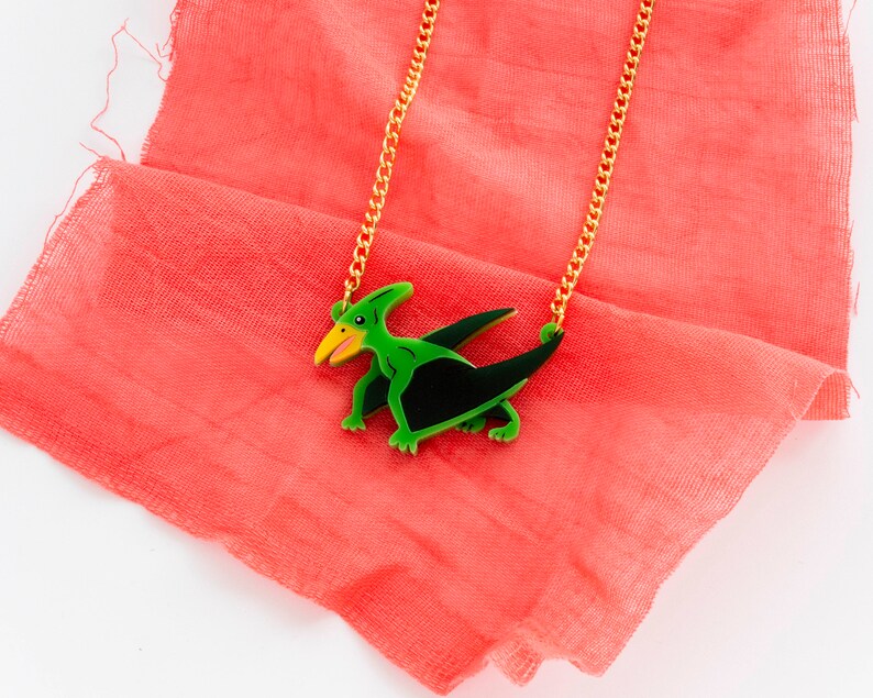 May include: A gold chain necklace with a green and black pterodactyl pendant. The pendant is made of wood and has a detailed design.
