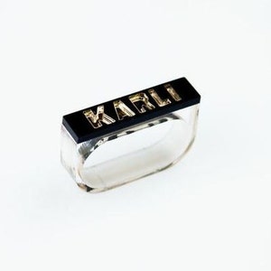 Puede incluir: Un anillo de acrílico transparente con un inserto rectangular negro que dice "KARLI" en letras doradas.