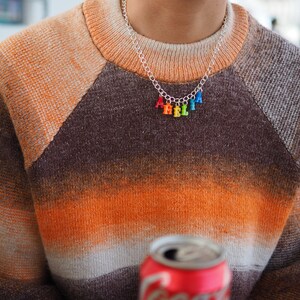 Puede incluir: Una persona lleva un su&eacute;ter a rayas marr&oacute;n, naranja y blanco con un collar de cadena plateada con abalorios de letras de colores que deletrean "HELLO".