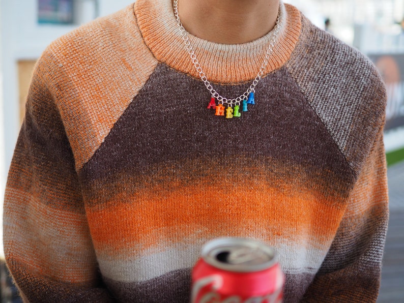 Puede incluir: Una persona lleva un su&eacute;ter a rayas marr&oacute;n, naranja y blanco con un collar de cadena plateada con abalorios de letras de colores que deletrean "HELLO".