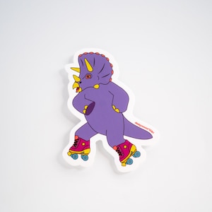 Könnte beinhalten: Lilafarbener Dinosaurier mit Rollschuhen mit gelben Rädern und rosa Schuhen. Der Dinosaurier hat gelbe Hörner und ein rosa-gelbes Design auf seinem Rücken. Der Dinosaurier befindet sich auf einem weißen Hintergrund.