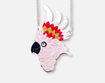 May include: A pink and white parrot pendant necklace with a silver chain. The parrot has a black beak, a white eye, and a crest with red, gold, and white accents. The necklace is a unique piece of jewelry.
