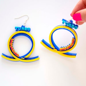 Pu&ograve; includere: Un paio di orecchini a forma di montagne russe blu e gialle con la scritta "TURBO" stampata sulla parte superiore. Le montagne russe sono in plastica e hanno un binario rosso.