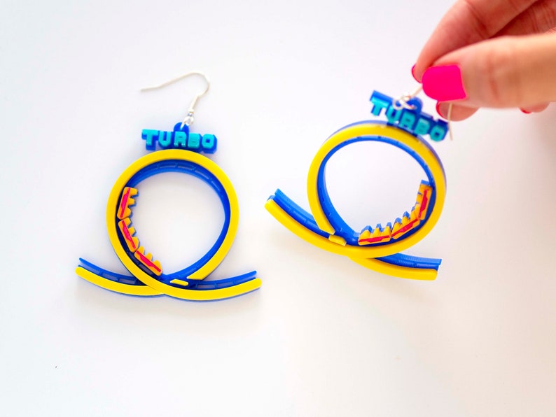 Pu&ograve; includere: Un paio di orecchini a forma di montagne russe blu e gialle con la scritta "TURBO" stampata sulla parte superiore. Le montagne russe sono in plastica e hanno un binario rosso.