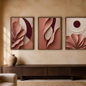 May include: A set of three framed wall art prints featuring abstract layered paper-like textures in shades of dusty rose, deep burgundy, and cream. The compositions include curved petal-like forms, circular shapes, and crescent arcs in a vertical orientation.