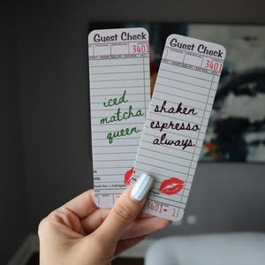 May include: Two rectangular paper bookmarks designed to resemble vintage guest checks. One features green cursive text reading iced matcha queen, while the other has black cursive text reading shaken espresso always. Both include red lip icons.