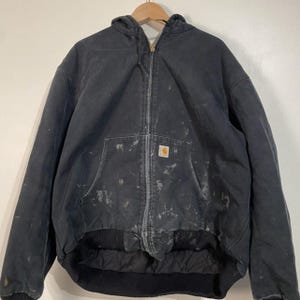 May include: A black zip-up hooded jacket featuring a front zipper closure, a drawstring hood, and a front pouch pocket. The fabric displays visible paint splatters throughout, and a small rectangular brand patch is sewn onto the front pocket.