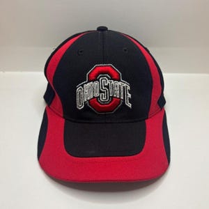 May include: A black and red baseball cap featuring the Ohio State logo embroidered in white and red on the front centre. The cap is constructed with a textured, breathable fabric and includes a structured crown and a curved brim in matching colours.