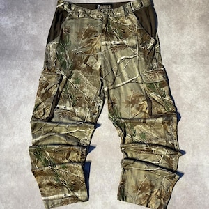 Könnte beinhalten: Cargohose mit Camouflage-Muster in Grün-, Braun- und Beigetönen. Die Hose verfügt über zwei große seitliche Cargotaschen mit Klappen, einen Knopf- und Reißverschluss an der Taille sowie ein schwarzes Etikett mit dem Schriftzug Rocky in der Mitte.