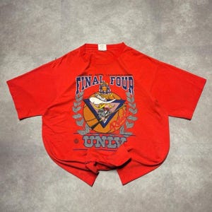 Puede incluir: Una camiseta roja de manga corta con un gráfico del torneo de baloncesto Final Four UNLV de 1991. El diseño incluye un personaje de dibujos animados con bigote y sombrero, un balón de baloncesto, hojas de laurel y el texto Final Four UNLV.
