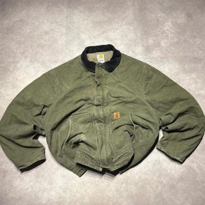 Può includere: Una giacca Carhartt verde oliva con colletto in velluto a coste nero, chiusura frontale con cerniera e patta con bottoni a pressione, e due ampie tasche anteriori. Una piccola etichetta quadrata Carhartt è visibile sulla tasca anteriore sinistra.