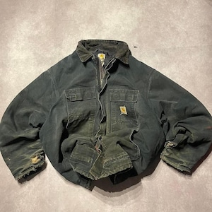 May include: A dark charcoal grey Carhartt work jacket featuring a fold-down collar, a full-length front zip, and two chest pockets with flaps. The jacket displays visible wear, including frayed cuffs and a small rectangular brand logo patch.