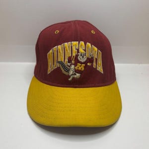 Könnte beinhalten: Eine kastanienbraune Baseballkappe mit gelbem Schirm. Auf der Vorderseite ist das Wort MINNESOTA in gelben Blockbuchstaben eingestickt, darunter befindet sich eine Grafik eines Zeichentrick-Gophers in einem kastanienbraunen und gelben Pullover.