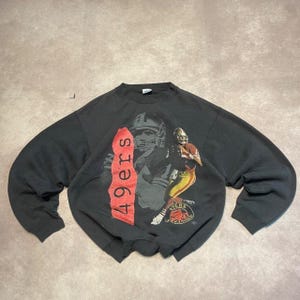 May include: A charcoal grey crewneck sweatshirt featuring a graphic print of a football player in a red and gold uniform. A vertical red brushstroke design contains the text 49ers in black, and a circular 49ers logo is positioned below.