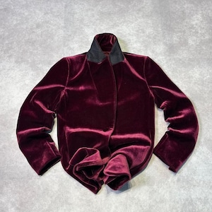 Puede incluir: Una chaqueta de manga larga confeccionada en tela de terciopelo color burdeos intenso. La prenda presenta un cuello estructurado en un material negro que contrasta, una tapeta frontal oculta y una textura suave y reflectante en toda la superficie.