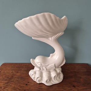 May include: A white ceramic decorative bowl shaped like a seashell supported by a sculpted dolphin base. The dolphin is depicted emerging from stylised waves, with its tail curving upwards to hold the shell-shaped basin in a smooth, glossy finish.