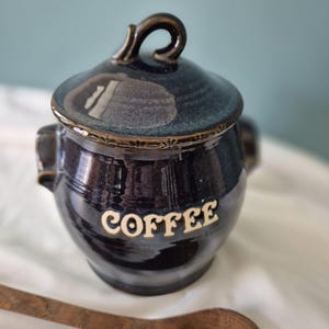 May include: A dark blue ceramic coffee canister with a matching lid featuring a curved handle and two side handles. The word COFFEE is embossed in cream-coloured lettering on the front. The glaze has a glossy finish with subtle variations.