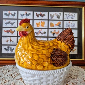 May include: A ceramic chicken-shaped lid sits atop a white, round, basket-weave patterned bowl. The chicken is primarily yellow with a red comb and wattle, featuring brown-painted wing and tail feathers with a glossy, textured finish.