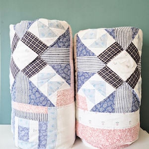 May include: Two rolled quilts featuring a patchwork design with various fabric patterns including blue and white stripes, dark navy plaid, light blue paisley, and soft pink floral prints. The textiles are stitched together in geometric blocks.