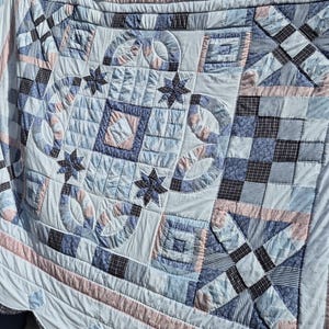May include: A patchwork quilt featuring a central square design with four dark blue star motifs and curved segments. The quilt is constructed from various fabric squares in shades of light blue, navy blue, white, and soft pink, with intricate stitching.