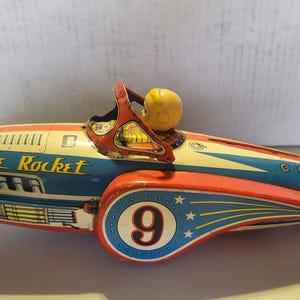 May include: A vintage tin toy space rocket in red, blue, and cream colors. The rocket features the text Space Rocket on the side, a large number 9 inside a blue circle with white stars, and a small yellow figure seated in the open cockpit area.