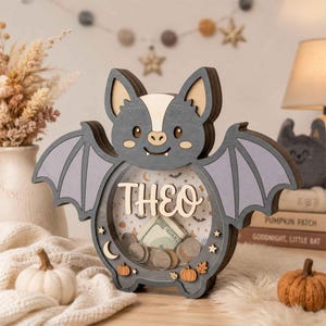 Puede incluir: Una hucha de madera con forma de murciélago y un panel frontal transparente que muestra el nombre THEO en letras en relieve. El interior presenta un fondo estampado con pequeños murciélagos, lunas y calabazas, conteniendo varias monedas.